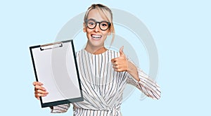 Beautiful blonde business woman holding clipboard with blank space smiling happy and positive, thumb up doing excellent and