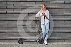Beautiful blonde in casual clothes standing with electric schooter outdoors at sunny daytime with phone in hands