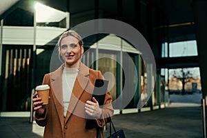 Beautiful blonde caucasian business woman walking to work carrying coffee and notebook