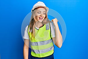 Beautiful blonde caucasian woman wearing architect hardhat pointing thumb up to the side smiling happy with open mouth