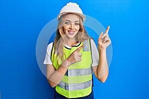 Beautiful blonde caucasian woman wearing architect hardhat smiling and looking at the camera pointing with two hands and fingers