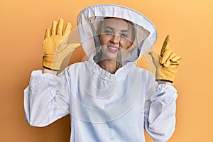 Beautiful blonde caucasian woman wearing protective beekeeper uniform showing and pointing up with fingers number seven while