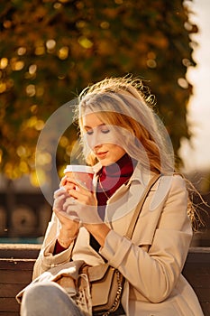 a beautiful blonde with a cup of coffee sits on a bench in an autumn park.