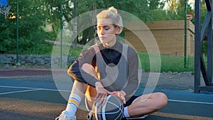 Beautiful blonde female basketball player sitting on ground and playing with ball, park in background