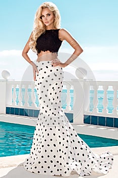 Beautiful blonde female model standing next to an exclusive pool in a long wedding dress