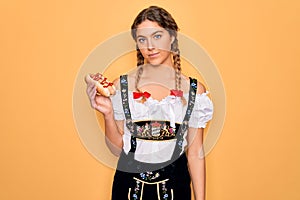 Beautiful blonde german woman with blue eyes wearing oktoberfest dress eating hot dog with a confident expression on smart face