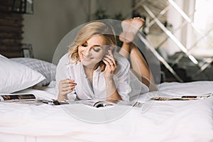 Beautiful blonde girl in bed lies on her stomach with her legs crossed listening to music through headphones and smiling