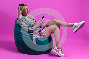 Beautiful blonde girl in a blue jacket and a purple sundress sits on a green bag chair with her cross leg on a pink background and