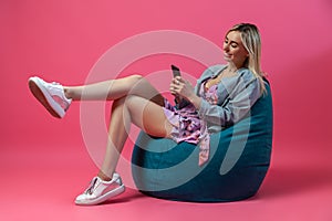 Beautiful blonde girl in a blue jacket and a purple sundress sits on a green bag chair with her cross leg on a pink background and photo