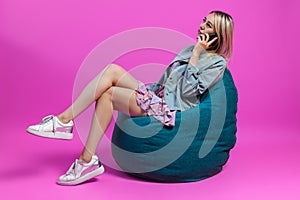 Beautiful blonde girl in a blue jacket and a purple sundress sits on a green bag chair with her legs folded on a pink isolated