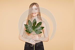 Beautiful blonde girl with clean skin holds green leaf on beige background. Beauty spa care cream concept