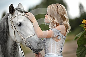 Beautiful blonde girl in dress strokes a gray horse on nature in