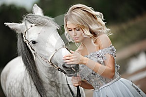 Beautiful blonde girl in dress strokes a gray horse on nature in