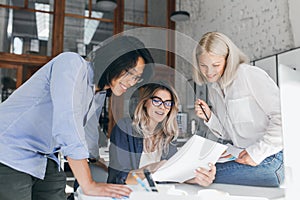 Beautiful blonde girl in glasses showing report to asian colleague which standing beside her table. Indoor portrait of