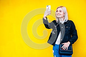 Beautiful blonde girl in headphones and smartphone in hands is talking on the phone through headphones on yellow wall background.