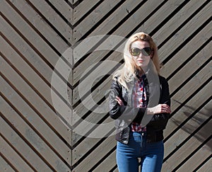 Beautiful blonde girl in huge sunglasses and a black jacket posing nex to wooden wall on a sunny day