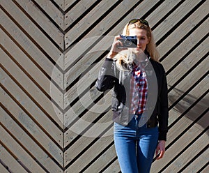 Beautiful blonde girl in huge sunglasses and a black jacket posing nex to wooden wall sunny day makes shoot vintage camera