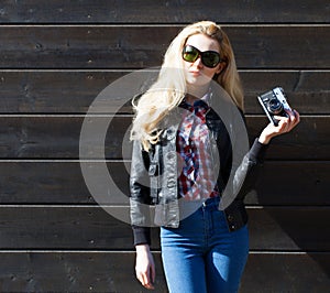 Beautiful blonde girl in huge sunglasses and a black jacket posing nex to wooden wall on a sunny day with a vintage camera