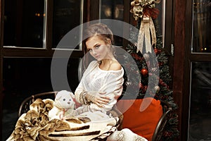 Beautiful blonde girl hugging herself sitting relaxing with warm blanket on sofa near Christmas tree and dark window