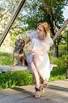 A beautiful blonde girl in a light dress with her cute dog is sitting on a park bench. Vertical view