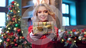 A beautiful blonde girl in a red dress stretches a gift box into the camera as a sign of the New year