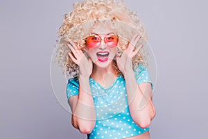 Beautiful blonde girl in retro style with voluminous curly hairstyle in a blue polka-dot blouse and pink glasses on a gray