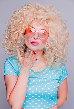 Beautiful blonde girl in retro style with a voluminous curly hairstyle, in a blue polka-dot blouse and pink glasses on a gray