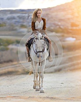 Beautiful blonde girl riding white horse in the mountain desert of Cappadocia in the rays of sunset