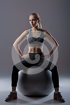Beautiful blonde girl in sports clothes sitting on fitness ball on dark background