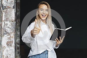 Beautiful blonde girl is standing in white shirt on dark background near brick wall in office, shows approving gesture.