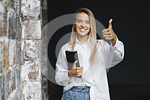 Beautiful blonde girl is standing in white shirt on dark background near brick wall in office, shows approving gesture. photo