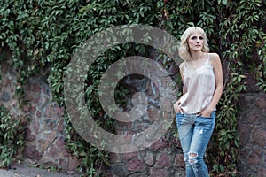 Beautiful blonde girl in a vest and jeans stands near wall with vine on city streets