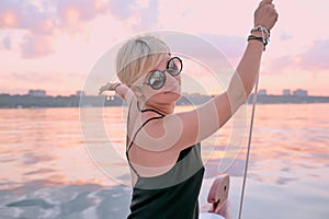 Beautiful blonde happy woman in sunglasses and long black dress enjoying sunset on the yacht in the sea.