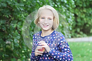 Beautiful blonde kid girl on a walk in the park eats fruit, nectarine or peach.