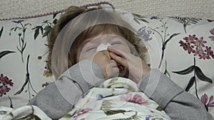 Beautiful blonde little girl lying in the bed, she is sick, but she smiling, blows her nose, tears her eyes, wipes