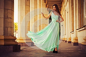 Beautiful blonde with a long curly hair in a long evening dress in static outdoors near retro vintage building in summer sunset