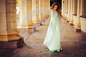 Beautiful blonde with a long curly hair in a long evening dress in static outdoors near retro vintage building in summer sunset