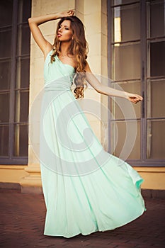 Beautiful blonde with a long curly hair in a long evening dress in static outdoors near retro vintage building in summer sunset