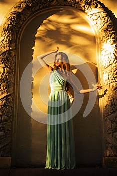 Beautiful blonde with a long curly hair in a long evening dress in static outdoors near retro vintage building in summer sunset