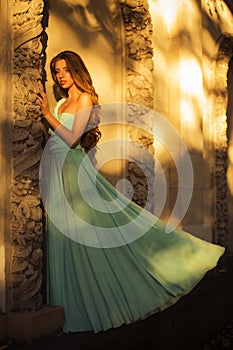 Beautiful blonde with a long curly hair in a long evening dress in static outdoors near retro vintage building in summer sunset