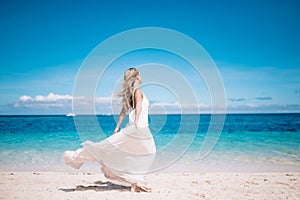 Beautiful blonde long hair bride in long white dress running on the white sand beach. Tropical turquois sea on the background.