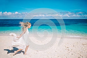 Beautiful blonde long hair bride in long white dress running on the white sand beach