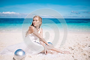 Beautiful blonde long hair bride in long white dress sitting on the white sand beach with a pearl