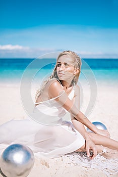 Beautiful blonde long hair bride in long white dress sitting on the white sand beach with a pearl