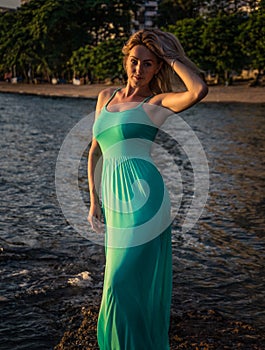 A beautiful blonde in a long turquoise dress stands on the seashore against a backdrop of a bright sunset