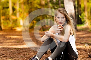 Beautiful blonde model in the middle of pines sitting using cellphone with headset. Listening to music