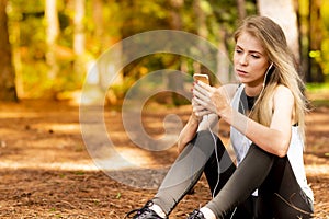 Beautiful blonde model in the middle of pines sitting using cellphone with headset. Listening to music