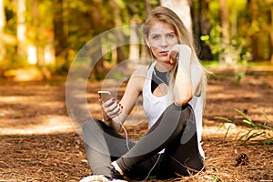 Beautiful blonde model in the middle of pines sitting using cellphone with headset. Listening to music