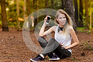 Beautiful blonde model in the middle of pines sitting using cellphone with headset. Listening to music