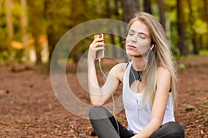 Beautiful blonde model in the middle of pines sitting using cellphone with headset. Listening to music